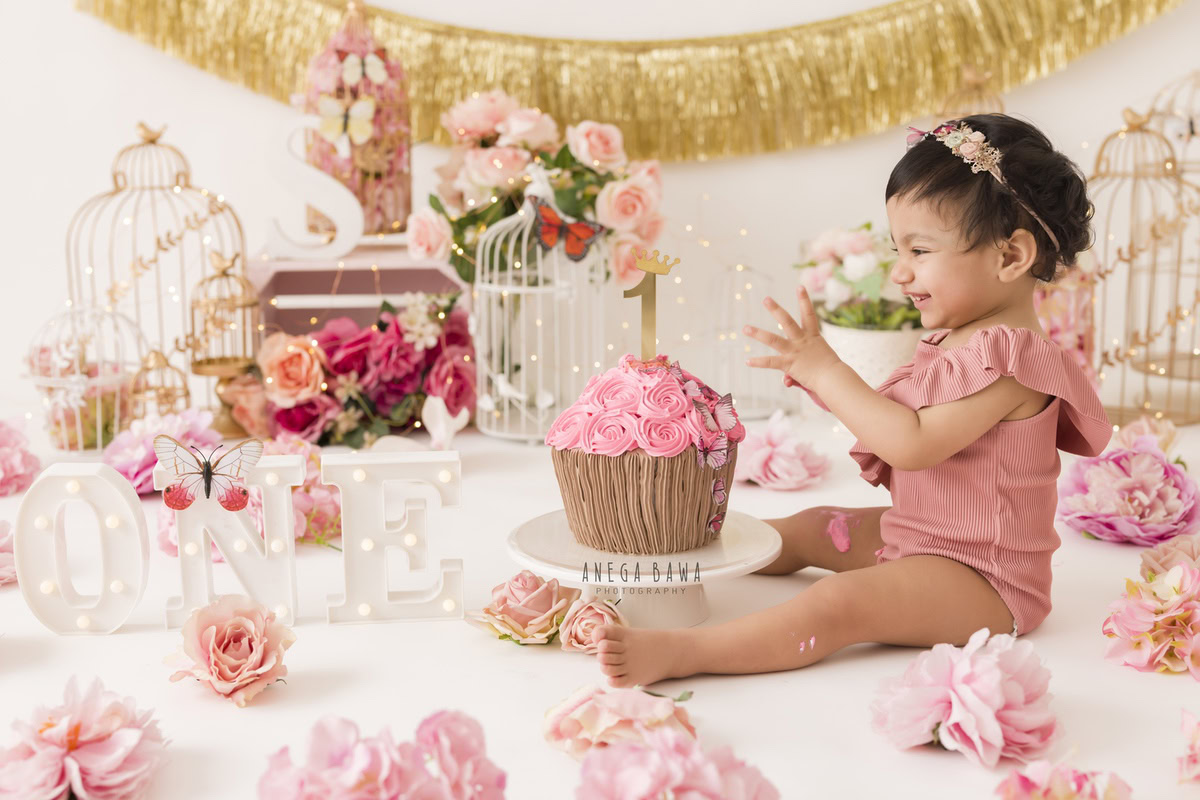 1 year old girl with a cake smash setup, featuring white castles, a golden fringe on the wall, and pink and peach flowers. Celebrating her first birthday. Cake Smash photo shoot in Delhi, Gurgaon by Anega Bawa Family Photographer Gurgaon (Delhi NCR).