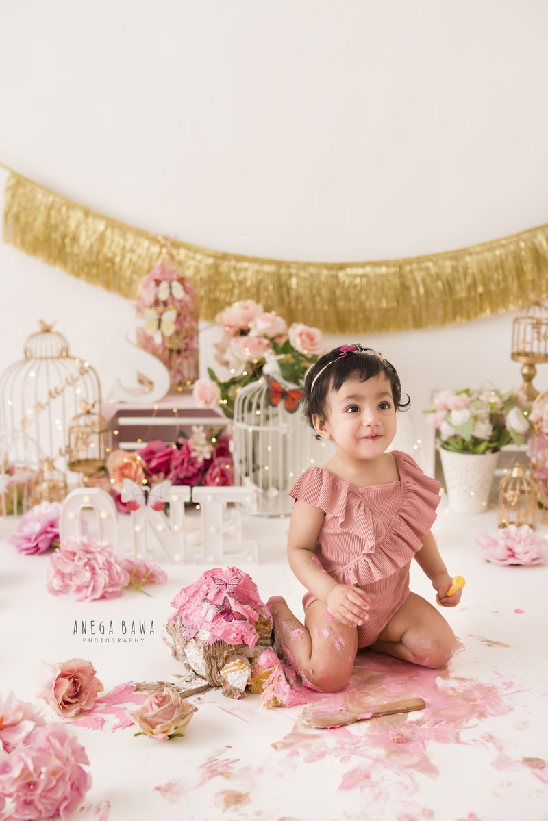1 year old girl with cake smash surrounded by white castles golden fringe on the wall pink and peach flowers and a white backdrop. Captured during a delightful first birthday photography session in Delhi Gurgaon by the renowned family photographer Anega Bawa.