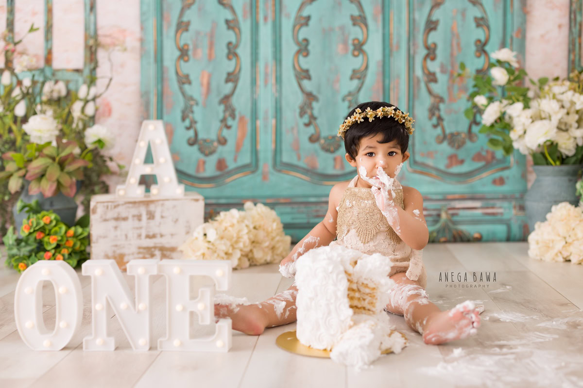 1 year old girl with a cake smash setup, featuring a green door backdrop, a green vase, and multicolored flowers. Celebrating her first birthday. Cake Smash photo shoot in Delhi, Gurgaon by Anega Bawa Family Photographer Gurgaon (Delhi NCR).