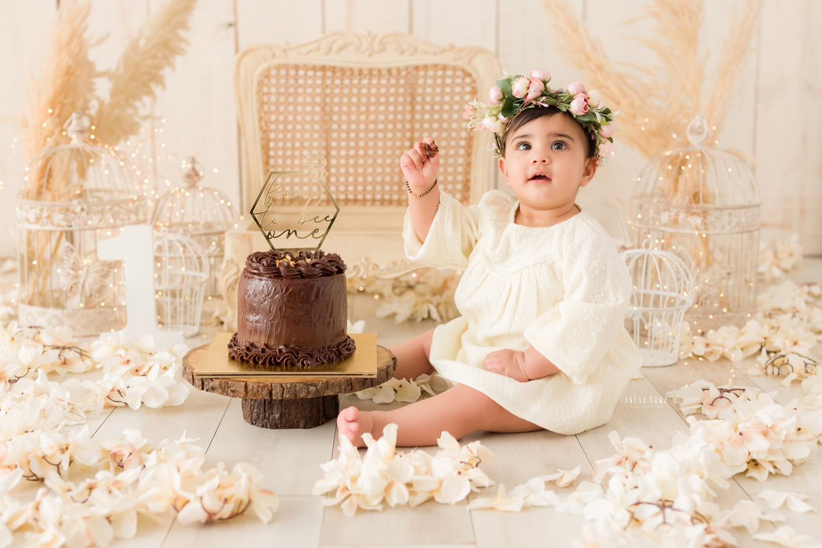 1year old girl with a cake smash setup featuring a wooden chair, white flowers, and white castles against a beige backdrop, captured by Anega Bawa during a Cake Smash photoshoot in Delhi, Gurgaon