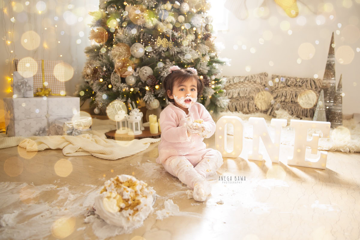 1 year old girl with cake smash surrounded by a Christmas tree fairy lights and an Eiffel Tower backdrop. Captured during a festive first birthday photo shoot in Delhi Gurgaon by the renowned family photographer Anega Bawa.