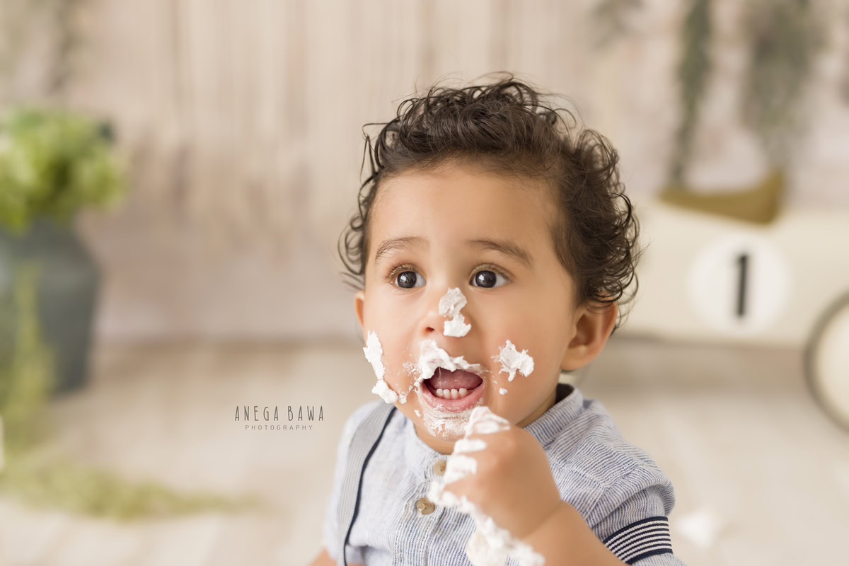 1 year old boy with cake smash on his face and curly hair against a beige backdrop. Captured during a delightful first birthday photoshoot in Delhi Gurgaon by the renowned family photographer Anega Bawa.