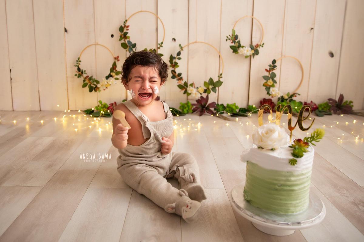 Boy with Cake Smash, beige backdrop, sparkling fairy lights, and a wooden floral frame on the wall, first birthday photo shoot in Delhi, Gurgaon. Captured by Anega Bawa family photographer Gurgaon (Delhi NCR)