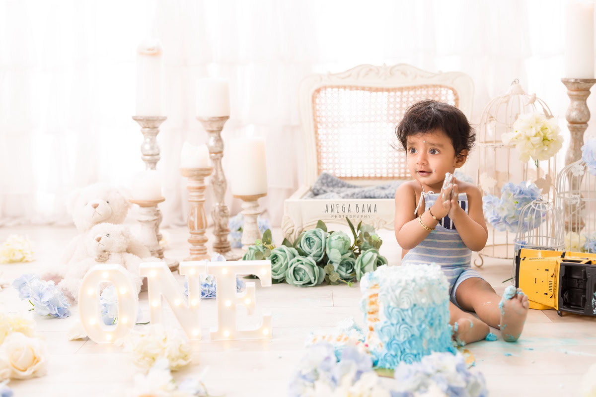 1year old boy with a cake smash setup featuring a candle stand, a white wooden chair, blue and white flowers, and teddy bears, captured by Anega Bawa during a Cake Smash photoshoot in Delhi, Gurgaon