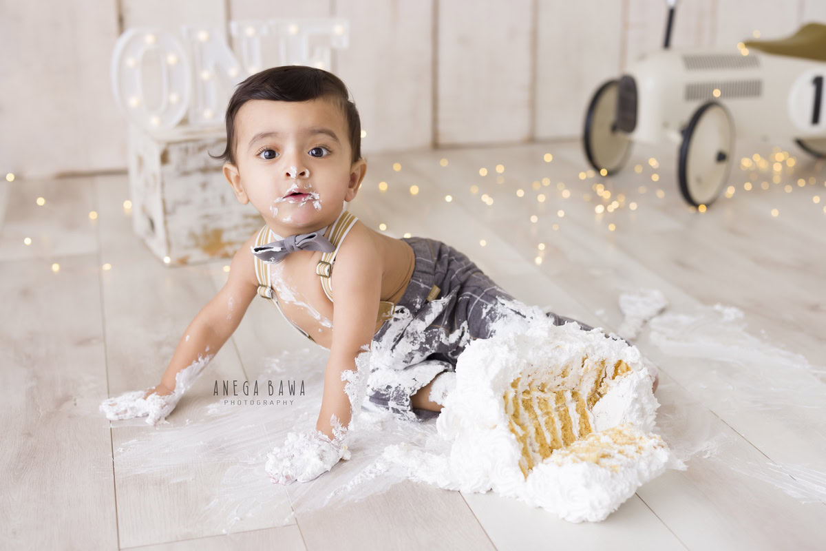 Boy enjoying cake smash with fairy lights, toy scooter, beige backdrop, first birthday photoshoot in Delhi, Gurgaon by Anega Bawa.