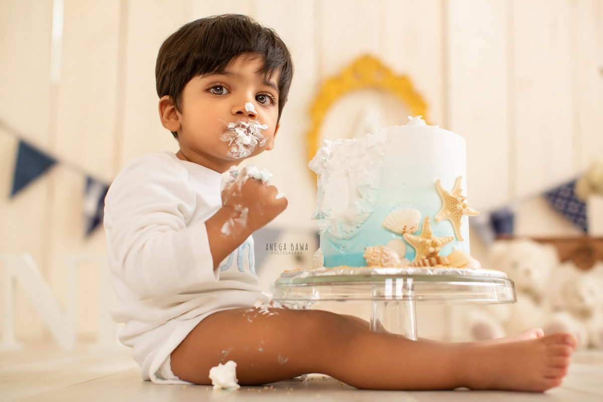 Boy with Cake Smash on face, beige backdrop, and a golden frame on the wall, first birthday photoshoot in Delhi, Gurgaon. Captured by Anega Bawa family photographer Gurgaon (Delhi NCR)
