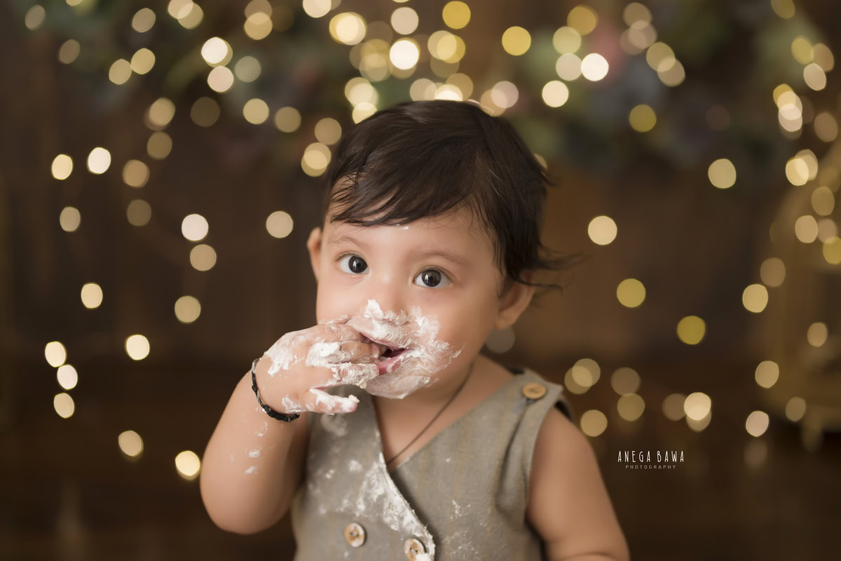 1-year-old boy with cake on his face and a cake smash setup against a brown backdrop with fairy lights, captured by Anega Bawa during a Cake Smash photoshoot in Delhi, Gurgaon