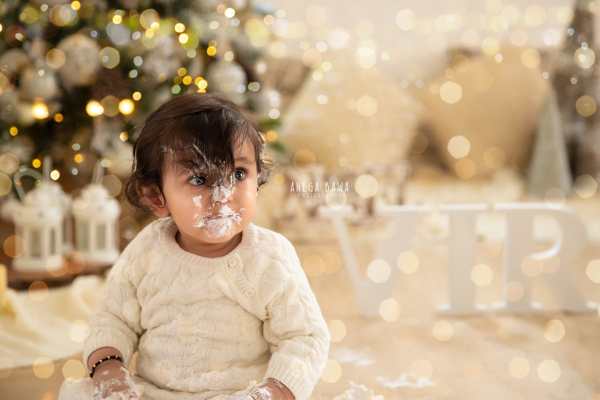 1 year old boy with cake smash on his face surrounded by fairy lights a Christmas tree and a name frame on the floor against a beige backdrop. Captured during a festive first birthday photography session in Delhi Gurgaon by the renowned family photographer Anega Bawa.