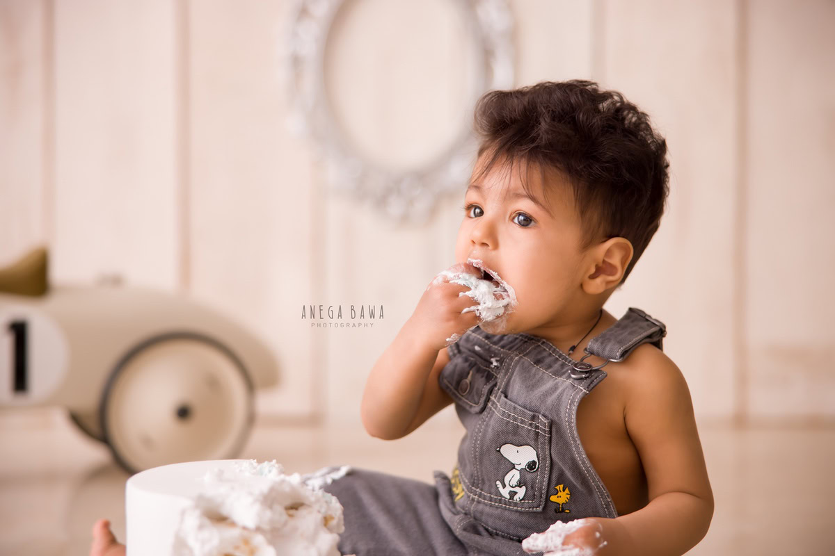 1 year old boy with cake smash on his face and a toy scooter against a beige backdrop. Captured during a charming first birthday photoshoot in Delhi Gurgaon by the renowned family photographer Anega Bawa.
