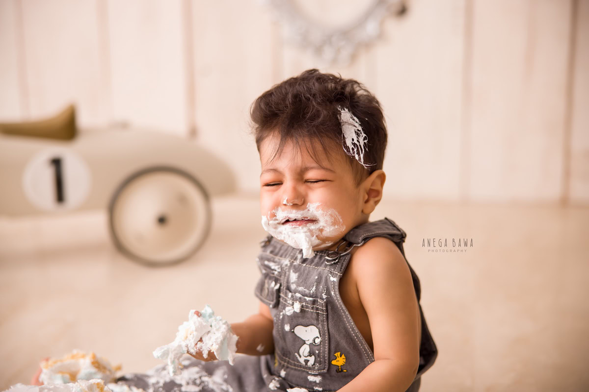 1 year old boy with cake smash on his face and a toy scooter against a beige backdrop. Captured during a delightful first birthday photoshoot in Delhi Gurgaon by the renowned family photographer Anega Bawa.