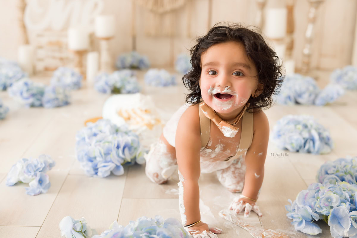 1year old boy crawling on the floor with cake on his face, surrounded by blue flowers and a candle stand, against a beige backdrop, captured by Anega Bawa during a Cake Smash photoshoot in Delhi, Gurgaon