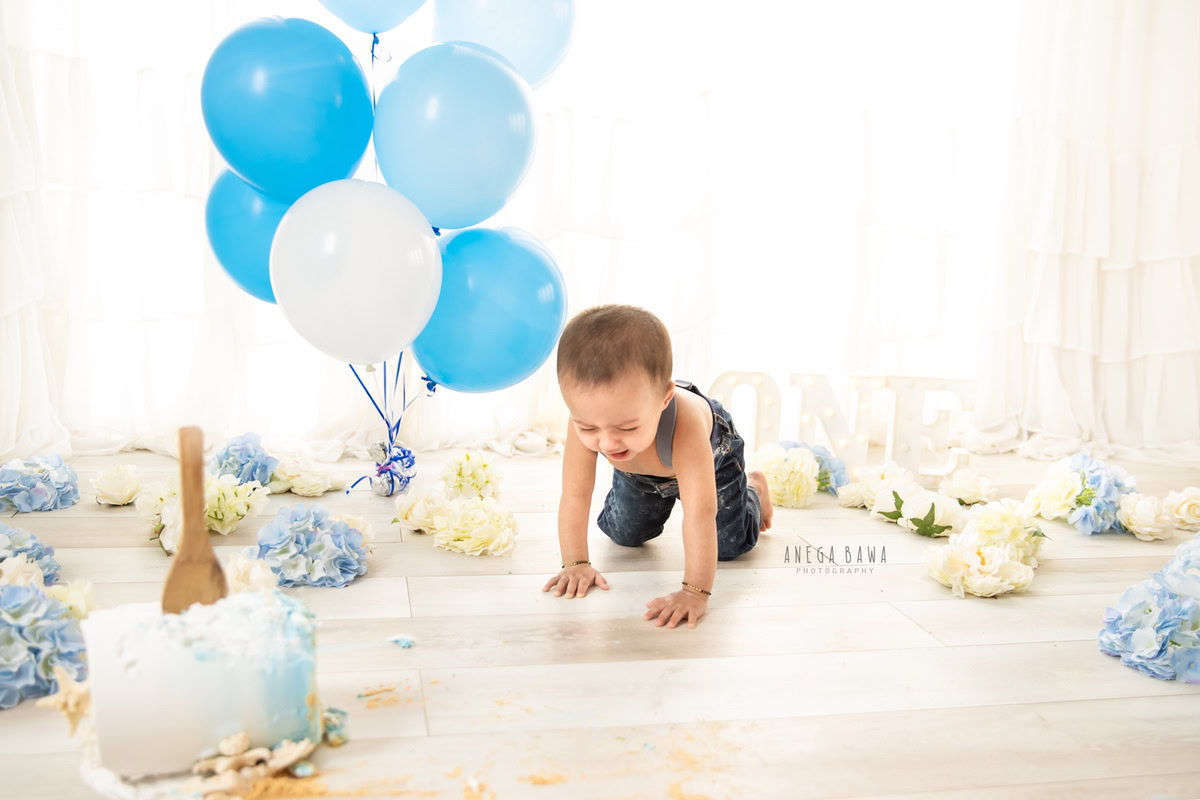 1 year old boy crawling on the floor with cake smash surrounded by blue and white balloons and flowers against a white backdrop. Captured during a delightful first birthday photoshoot in Delhi Gurgaon by the renowned family photographer Anega Bawa.