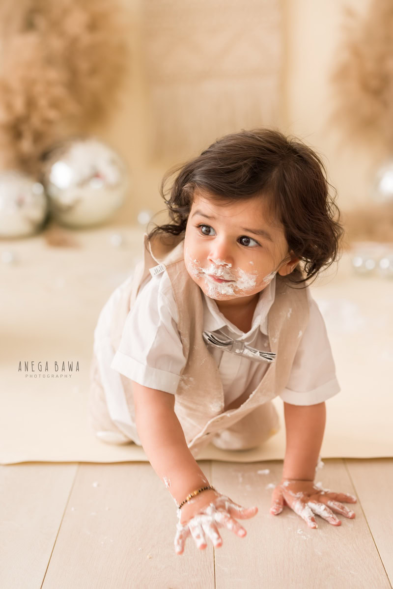 1year old boy crawling with cake on his face and a cake smash setup featuring bushes and disco balls against a beige backdrop, captured by Anega Bawa during a Cake Smash photoshoot in Delhi, Gurgaon