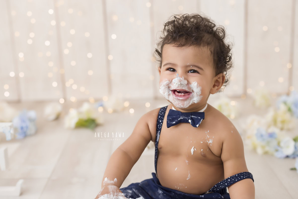 1 year old boy with a cute smile and cake smash on his face against a beige backdrop with fairy lights and flowers on the floor. Captured during a charming first birthday photoshoot in Delhi Gurgaon by the renowned family photographer Anega Bawa.