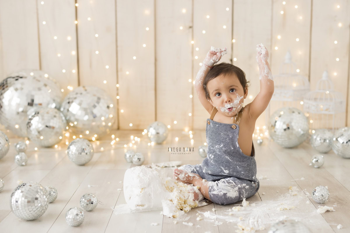 Boy in denim dungaree with Cake Smash photography featuring cake on face, surrounded by disco balls and sparkling fairy lights against a beige backdrop, first birthday photo shoot in Delhi, Gurgaon. Captured by Anega Bawa family photographer Gurgaon (Delhi NCR)