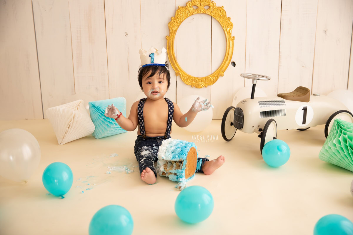 Boy in denim dungaree with Cake Smash, playing with a toy scooter, surrounded by blue balloons, pompom decorations, and a golden frame, against a beige backdrop, first birthday photoshoot in Delhi, Gurgaon. Captured by Anega Bawa family photographer Gurgaon (Delhi NCR)