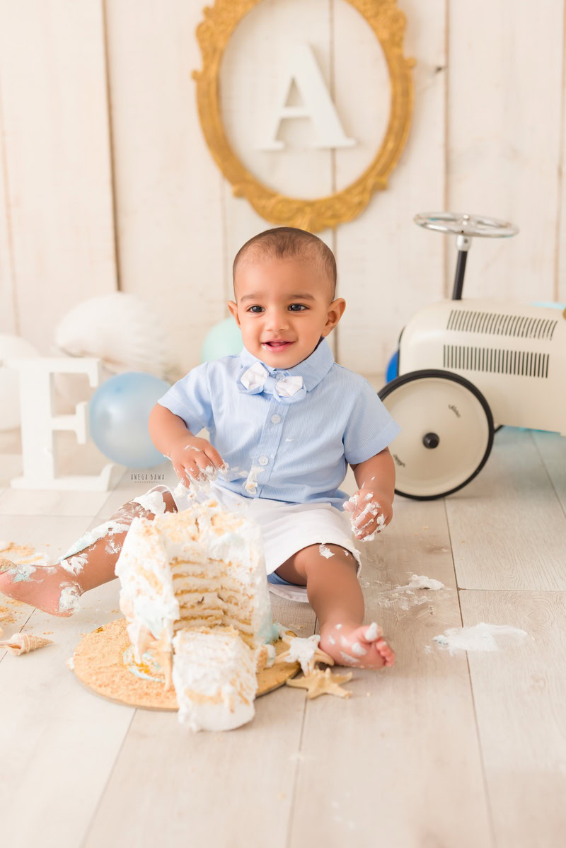 1 year boy enjoying a cake smash setup with a toy scooter and an alphabet frame on the wall against a beige backdrop, captured by Anega Bawa during a Cake Smash photoshoot in Delhi, Gurgaon