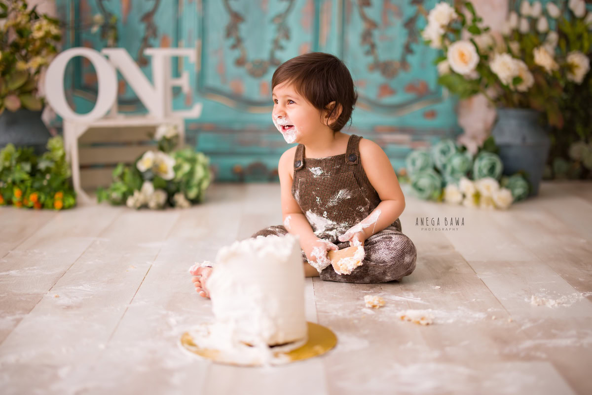 1 year old boy in brown dungarees with cake smash on his face against a green door backdrop and multicolour flowers. Captured during a delightful first birthday photography session in Delhi Gurgaon by the renowned family photographer Anega Bawa.