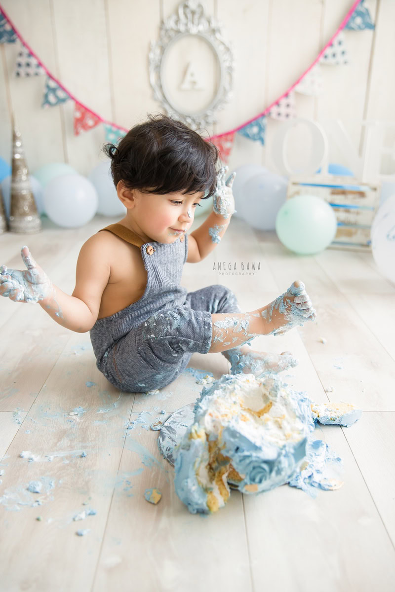 Boy playing with Cake Smash, surrounded by balloons, an alphabet silver frame on the wall, and a beige backdrop, first birthday photo shoot in Delhi, Gurgaon. Captured by Anega Bawa family photographer Gurgaon (Delhi NCR)