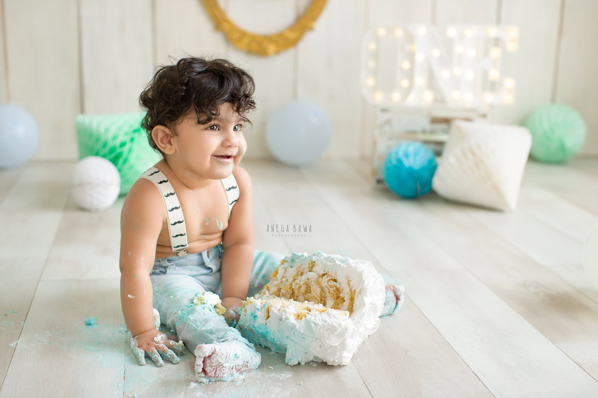 1 year old boy playing with cake smash against a beige backdrop with blue and green pompom balloons. Captured during a charming first birthday photography session in Delhi Gurgaon by the renowned family photographer Anega Bawa.