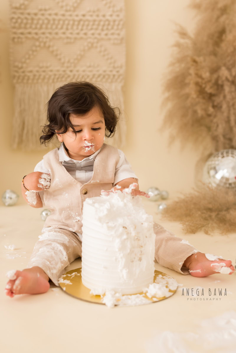 1 year boy playing with a cake smash setup against a beige backdrop with bushes and disco balls, captured by Anega Bawa during a Cake Smash photoshoot in Delhi, Gurgaon