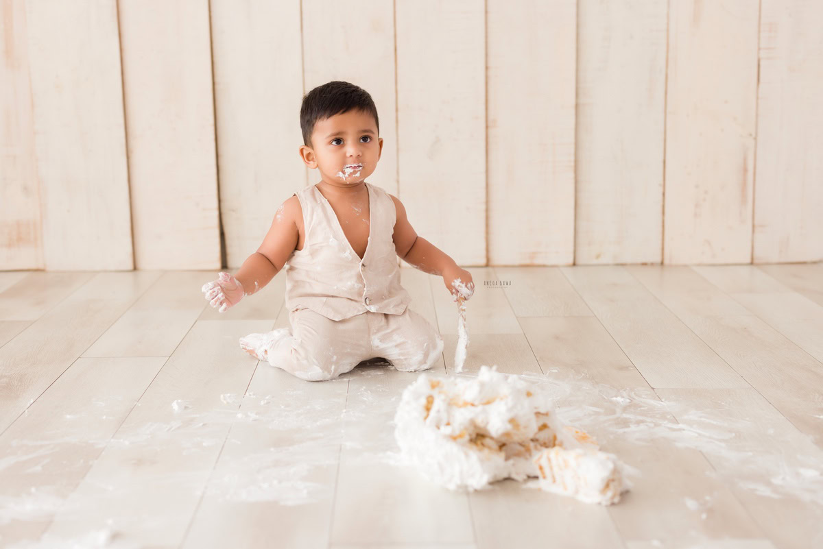 Boy playing with Cake Smash against a beige backdrop, first birthday photo shoot in Delhi, Gurgaon. Captured by Anega Bawa family photographer Gurgaon (Delhi NCR)