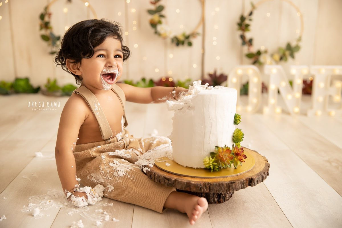 Boy playing with cake smash, beige backdrop, wooden leafy frame, charming first birthday photo shoot in Delhi, Gurgaon by Anega Bawa.