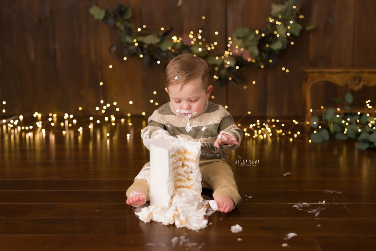 1 year boy playing with a cake smash setup against a brown backdrop with fairy lights, captured by Anega Bawa during a Cake Smash photoshoot in Delhi, Gurgaon