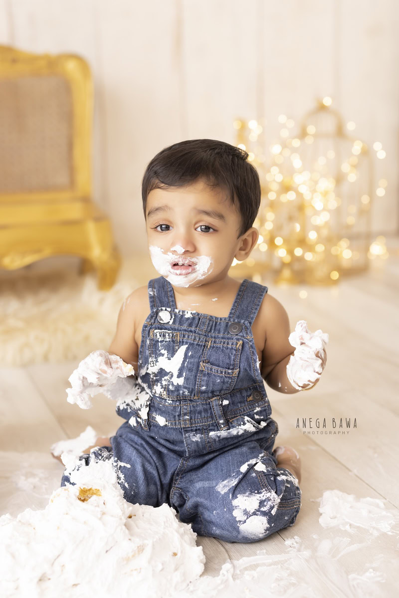 1 year boy playing with a cake smash setup featuring a golden chair, golden castles, and fairy lights against a beige backdrop, captured by Anega Bawa during a Cake Smash photoshoot in Delhi, Gurgaon