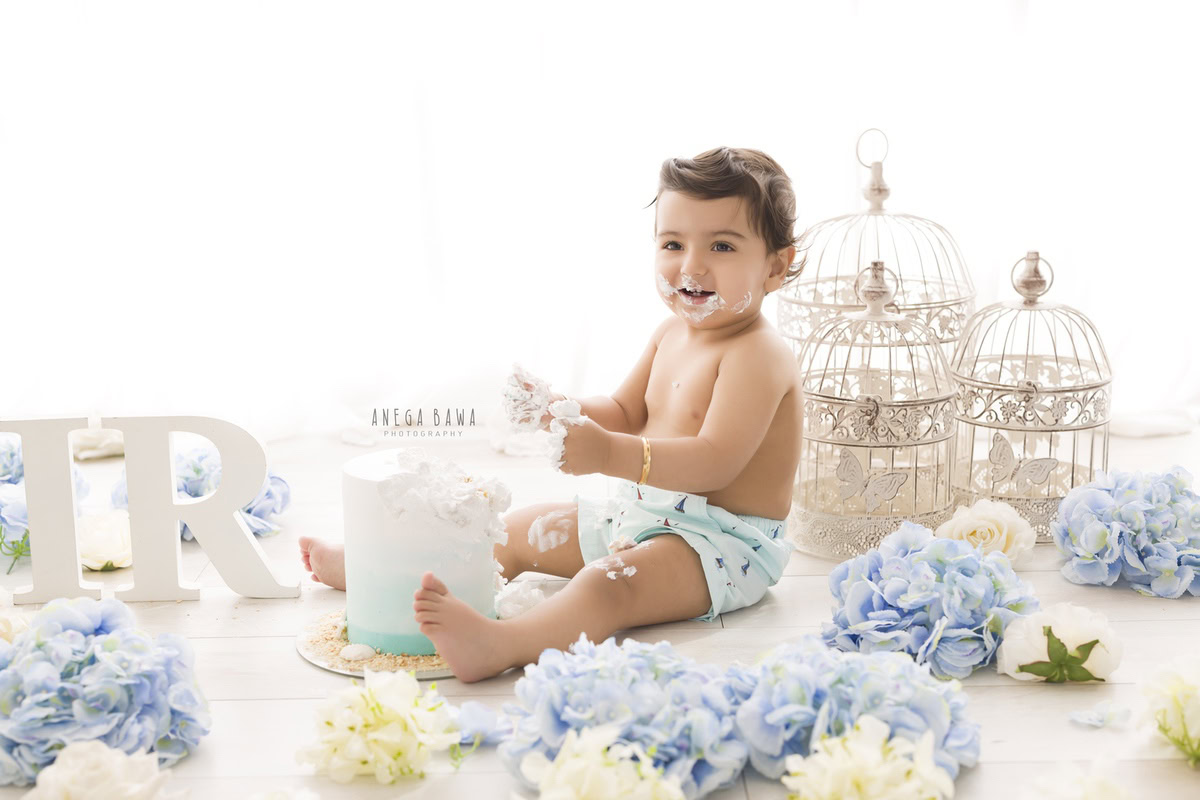 Boy playing with a cake during a Cake Smash photoshoot by Anega Bawa, set against a white backdrop with white castles, blue and white flowers in Delhi, Gurgaon.