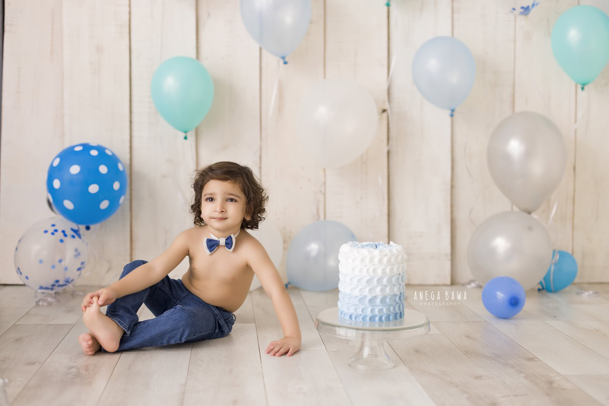 Boy posing with cake smash, wearing a cute bow, beige backdrop, multicolour balloons. First birthday photoshoot in Delhi, Gurgaon. Captured by Anega Bawa family photographer Gurgaon (Delhi NCR).