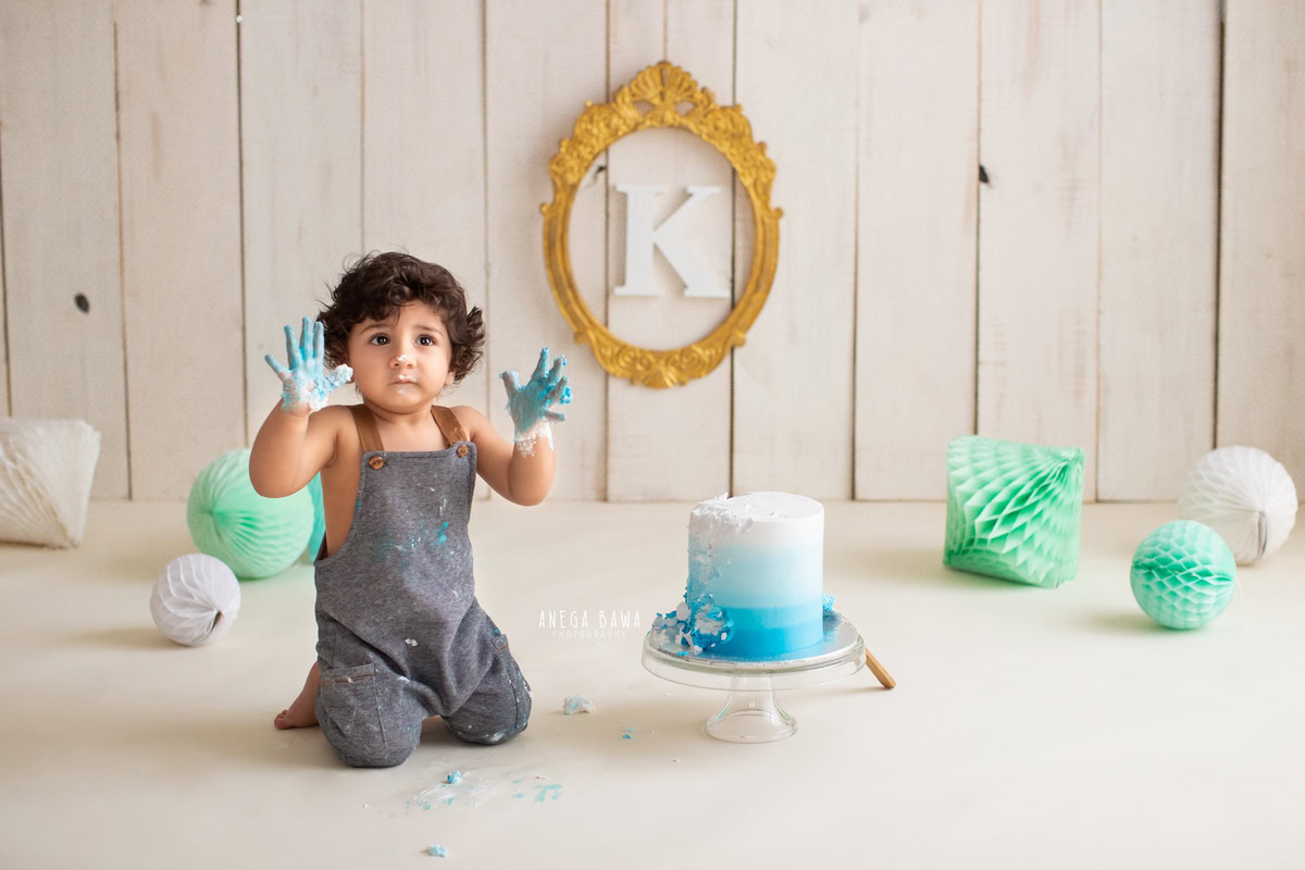 Boy posing with Cake Smash, featuring pompom decorations and an alphabet frame on the wall against a beige backdrop, first birthday photo shoot in Delhi, Gurgaon. Captured by Anega Bawa family photographer Gurgaon (Delhi NCR)