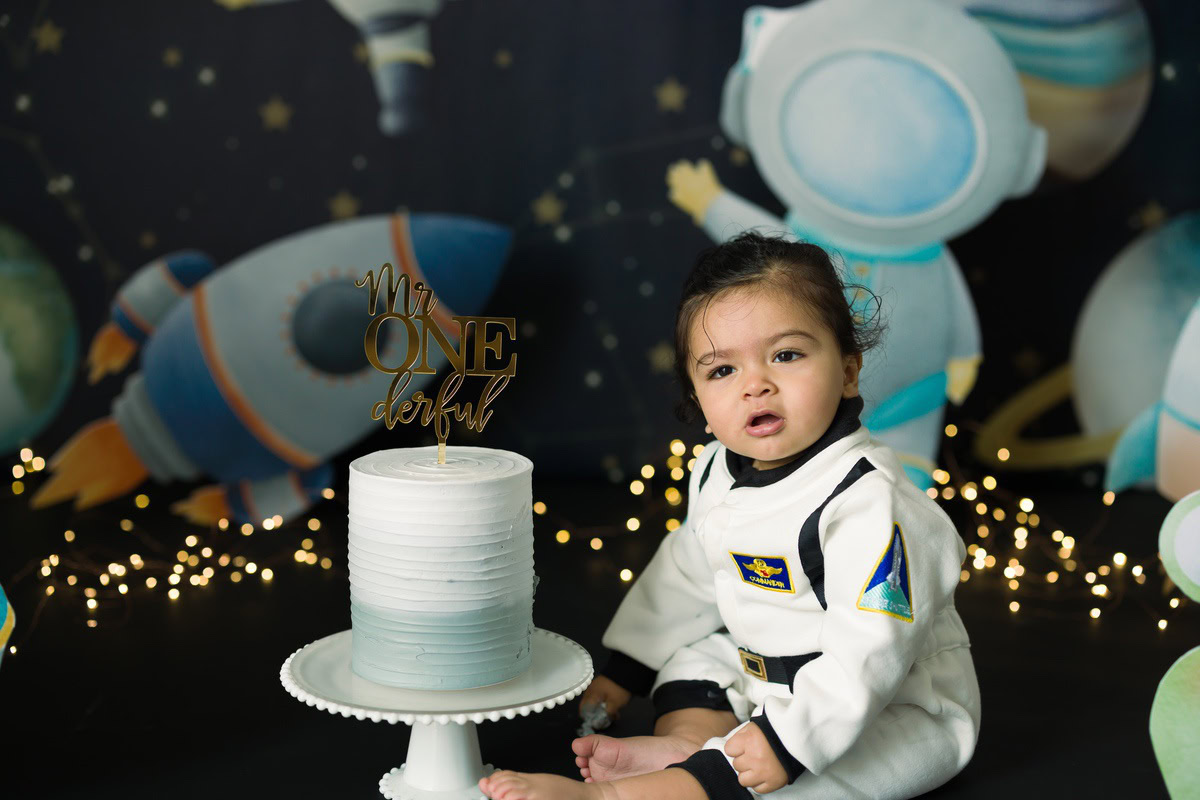 Boy posing with Cake Smash, surrounded by a toy spaceship and sparkling fairy lights, first birthday photoshoot in Delhi, Gurgaon. Captured by Anega Bawa family photographer Gurgaon (Delhi NCR)