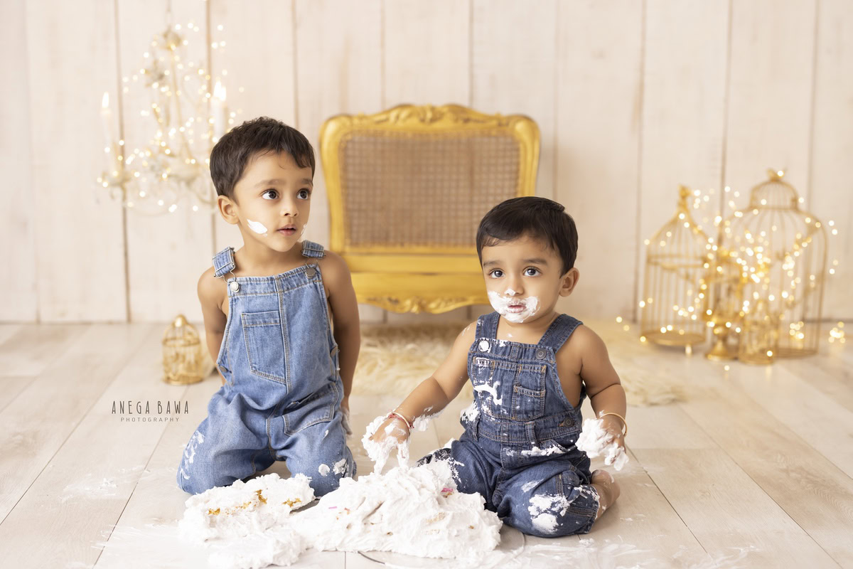 Boy sibling wearing denim dungaree, cake smash, golden chair, golden castles, candle stand, fairy lights, beige backdrop. First birthday photography in Delhi, Gurgaon. Captured by Anega Bawa family photographer Gurgaon (Delhi NCR).