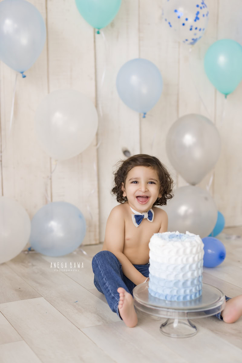 Boy sitting on the floor with a cake, surrounded by blue and silver balloons against a beige backdrop during a Cake Smash photoshoot by well-known Anega Bawa Photography in Delhi, Gurgaon.