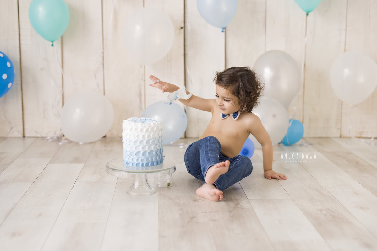 1 year old boy sitting on the floor with cake smash surrounded by multicolour balloons against a beige backdrop. Captured during a delightful first birthday photoshoot in Delhi Gurgaon by the renowned family photographer Anega Bawa.