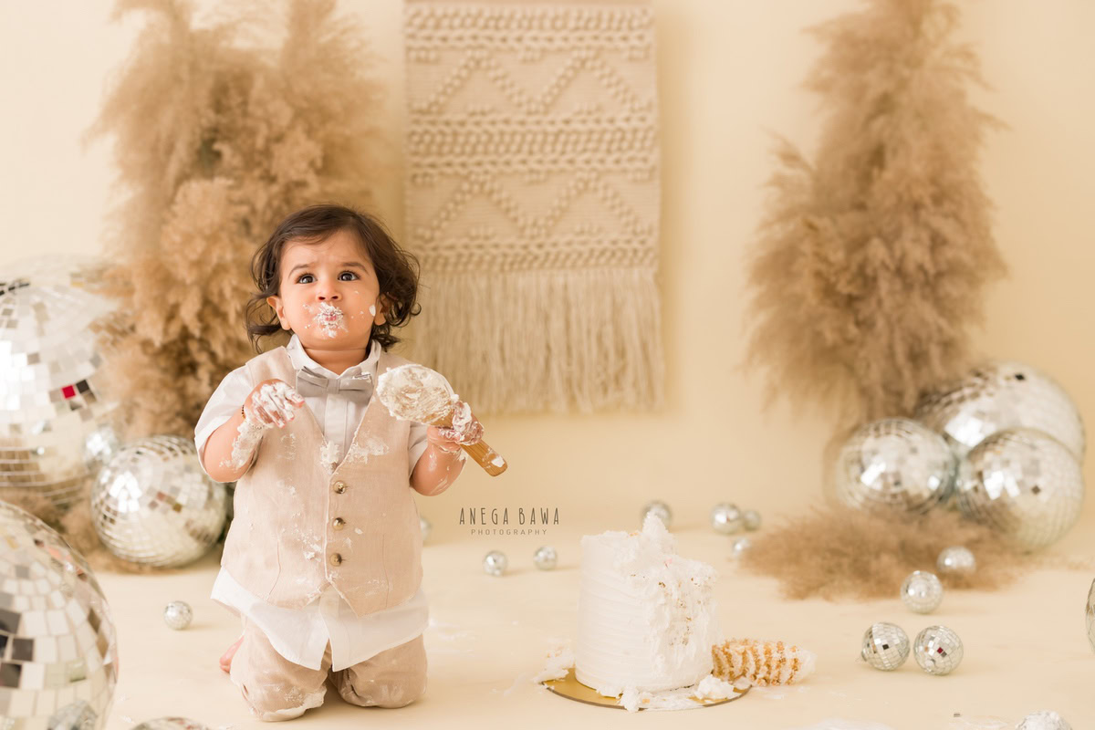 Boy sitting on floor with cake smash on his face, brown bushes, discoballs, beige backdrop. First birthday photoshoot in Delhi, Gurgaon. Captured by Anega Bawa family photographer Gurgaon (Delhi NCR).