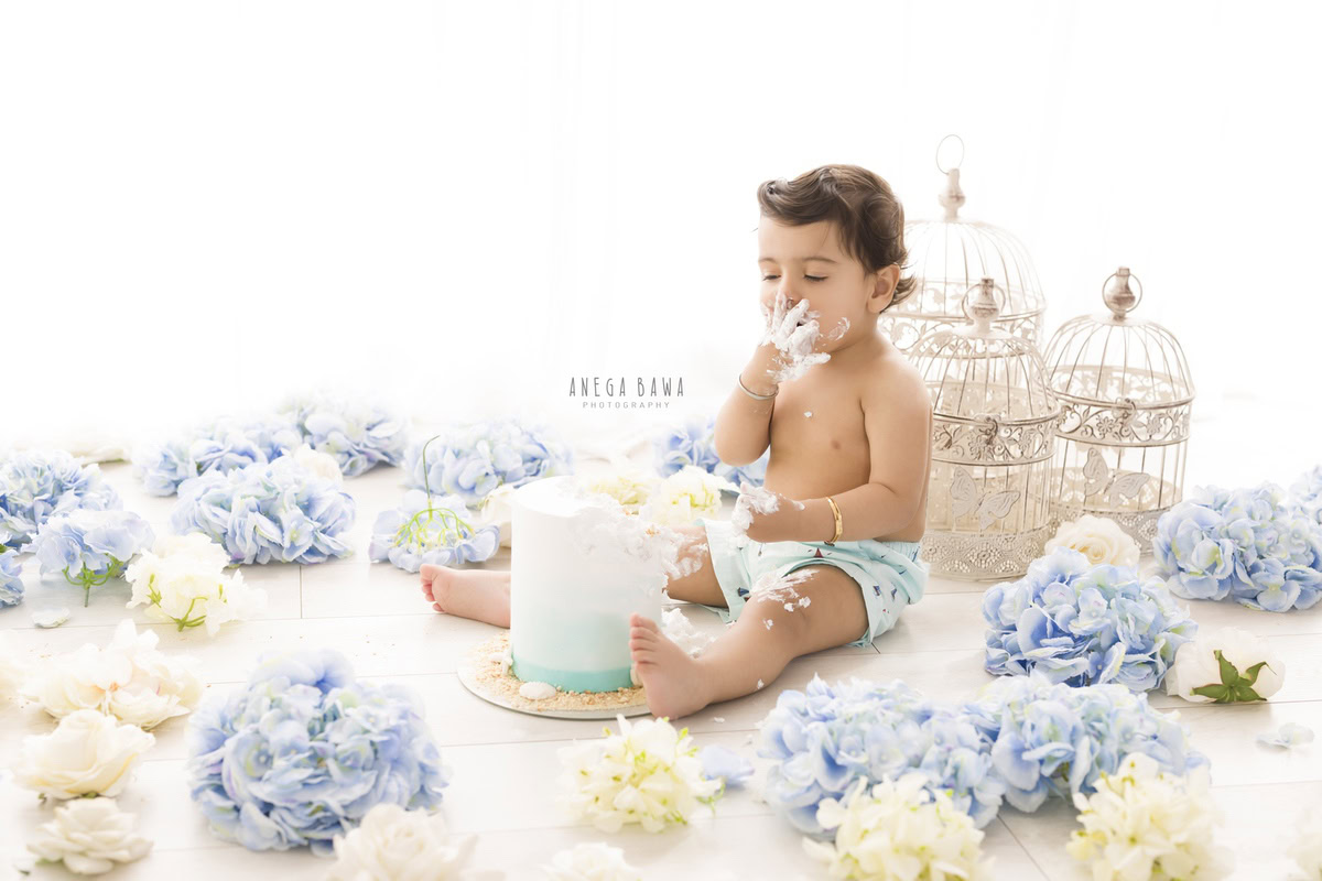 Boy sitting on the floor with a cake, surrounded by white castles, blue and white flowers against a white backdrop during a Cake Smash photoshoot by well-known Anega Bawa Photography in Delhi, Gurgaon.