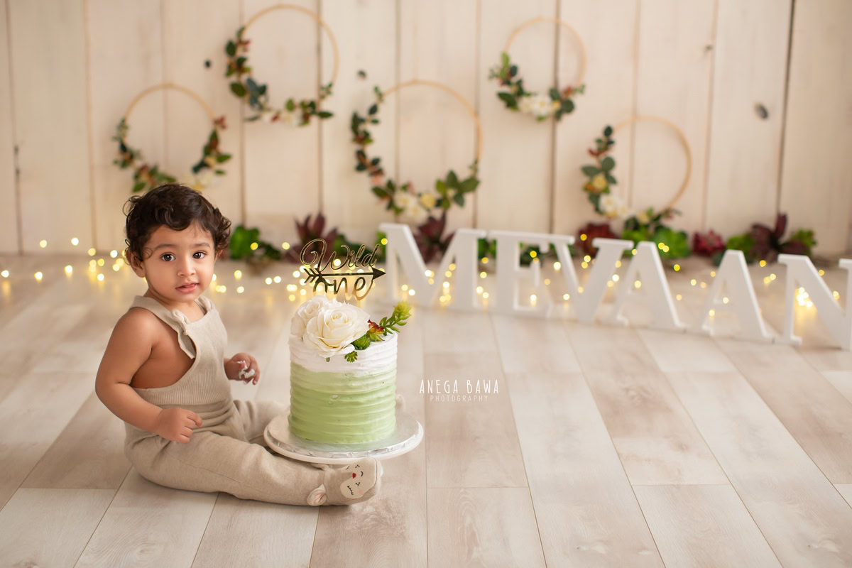 Boy sitting with cake smash on floor, name frame on floor, fairy lights, wooden floral frame, beige backdrop. First birthday photo shoot in Delhi, Gurgaon. Captured by Anega Bawa family photographer Gurgaon (Delhi NCR).