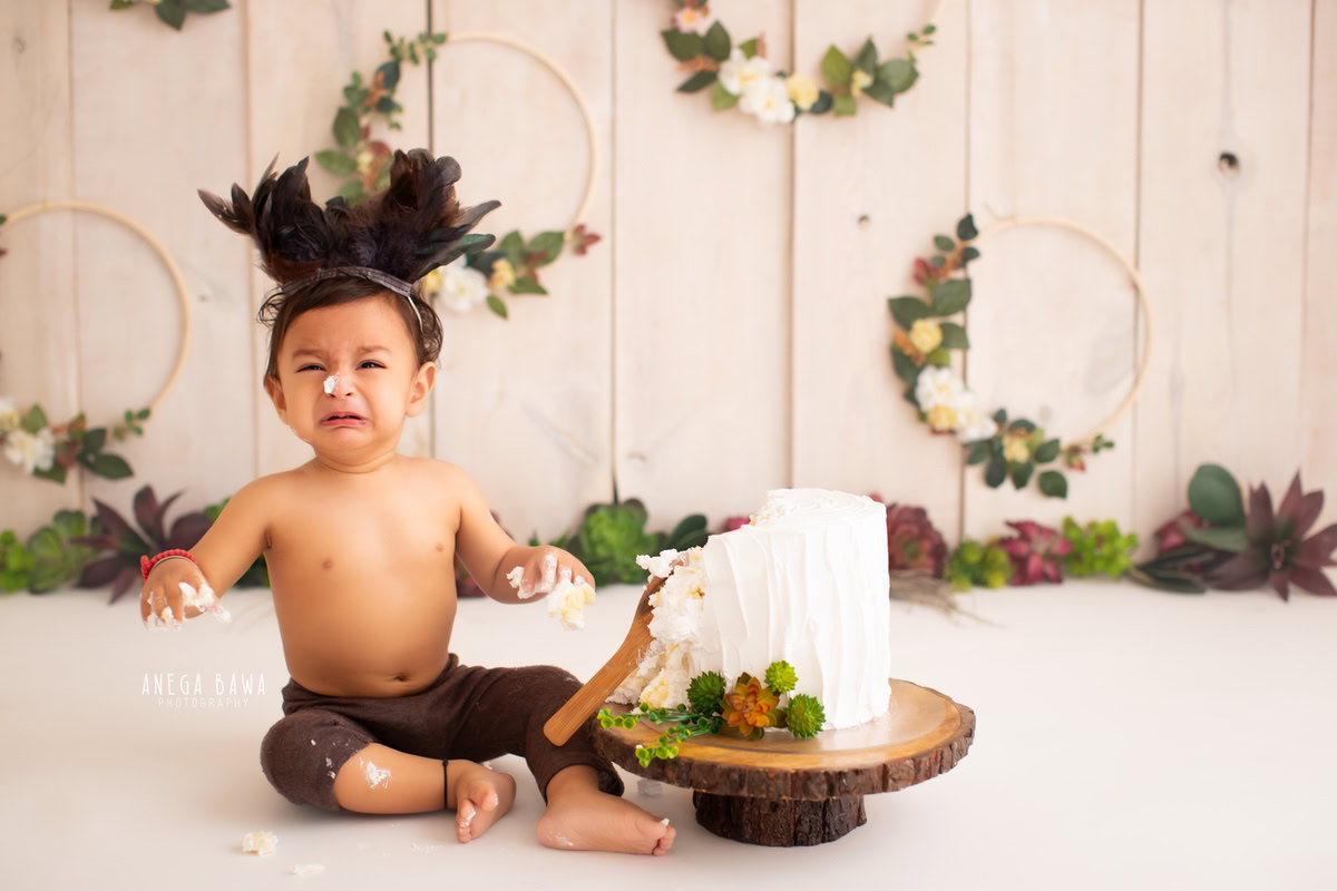 Boy sitting with cake smash, round wooden leafy frame on wall, beige backdrop. First birthday photoshoot in Delhi, Gurgaon. Captured by Anega Bawa family photographer Gurgaon (Delhi NCR).