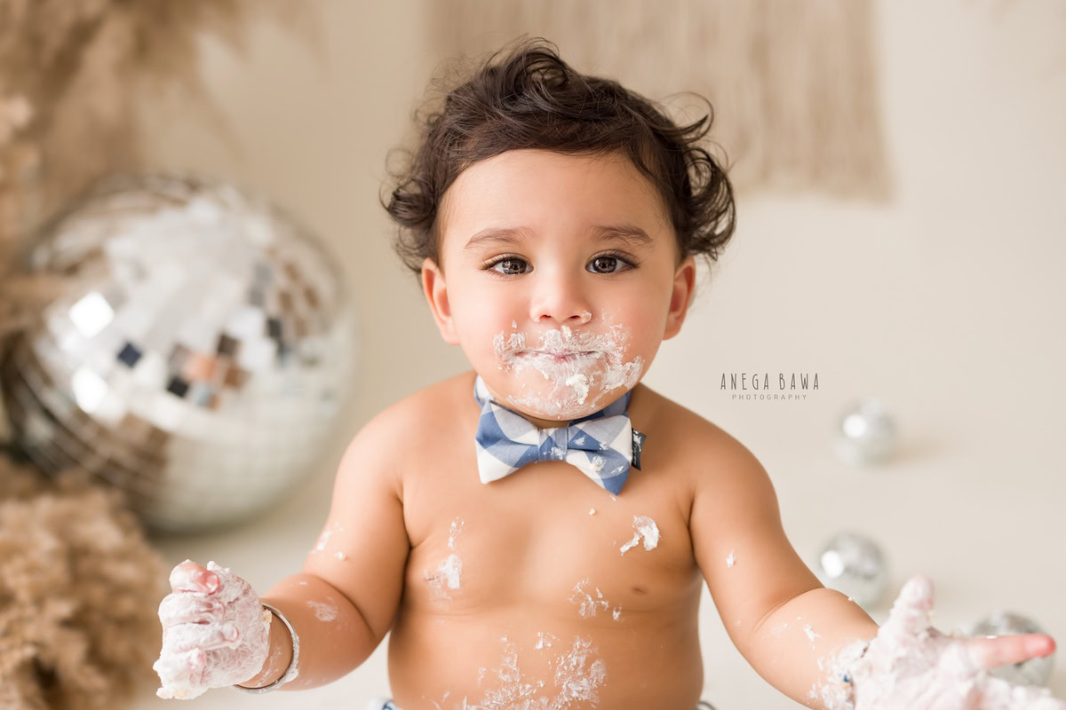Boy with cake smash on face, discoballs, bushes, beige backdrop. First birthday photography in Delhi, Gurgaon. Captured by Anega Bawa family photographer Gurgaon (Delhi NCR).