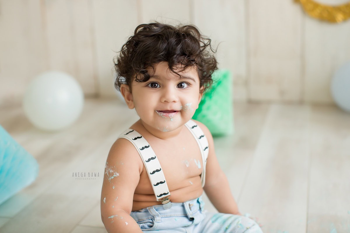 Boy with curly hair and Cake Smash on face, against a beige backdrop with blue and green pompom decorations, first birthday photoshoot in Delhi, Gurgaon. Captured by Anega Bawa family photographer Gurgaon (Delhi NCR)
