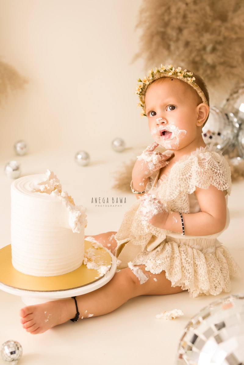 Girl with Cake Smash, wearing a cute headband, surrounded by bushes, beige backdrop, and sparkling disco balls, first birthday photography in Delhi, Gurgaon. Captured by Anega Bawa family photographer Gurgaon (Delhi NCR)