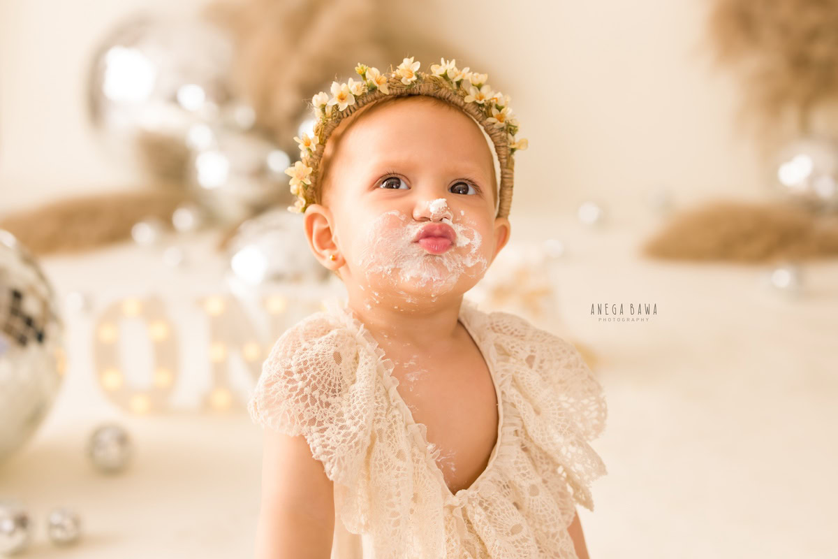 1 year old girl with cake smash on her face in a cute pout pose against a beige backdrop with discoballs and bushes. Captured during a charming first birthday photoshoot in Delhi Gurgaon by the renowned family photographer Anega Bawa.