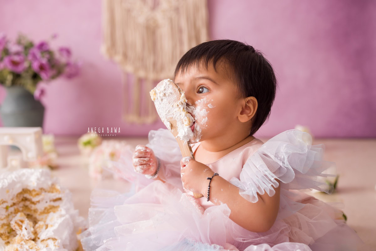 1 year old girl with cake smash on her face against a lavender backdrop with a green vase. Captured during a delightful first birthday photo shoot in Delhi Gurgaon by the renowned family photographer Anega Bawa.