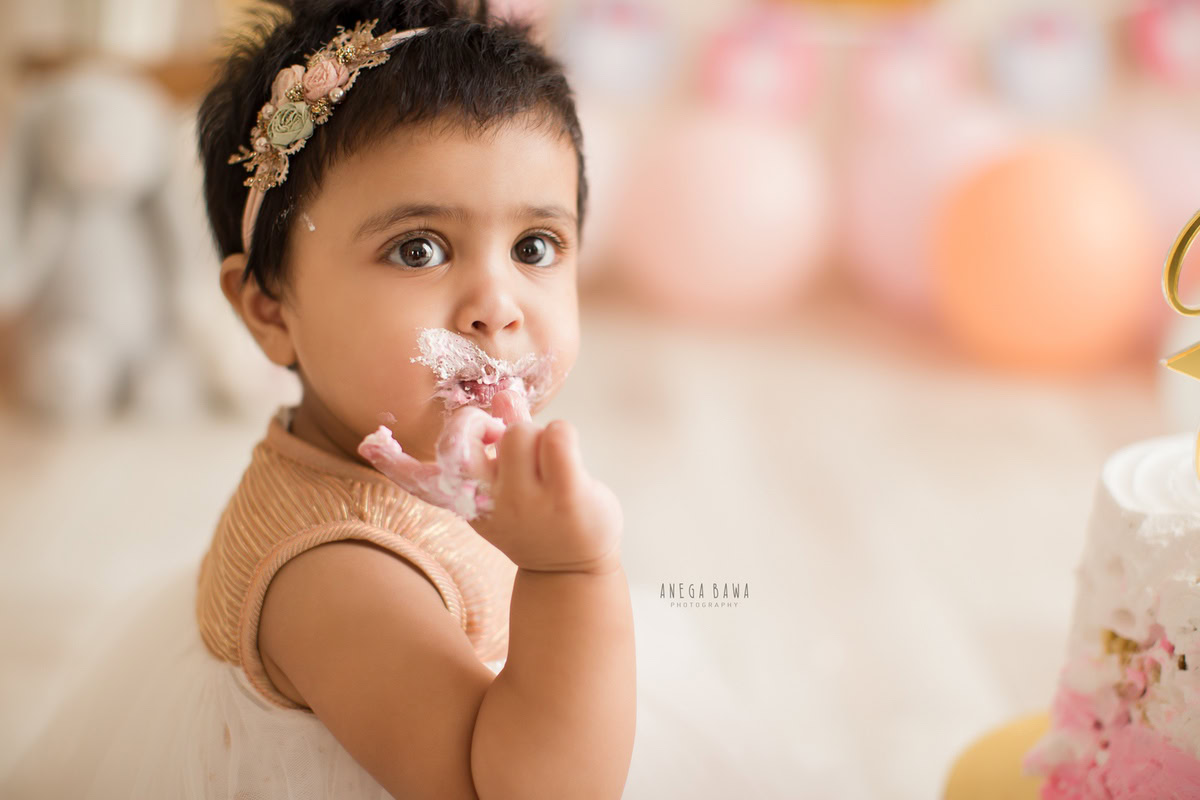 Girl with Cake Smash on face, surrounded by peach and pink balloons and cuddly teddy bears, first birthday photoshoot in Delhi, Gurgaon. Captured by Anega Bawa family photographer Gurgaon (Delhi NCR)