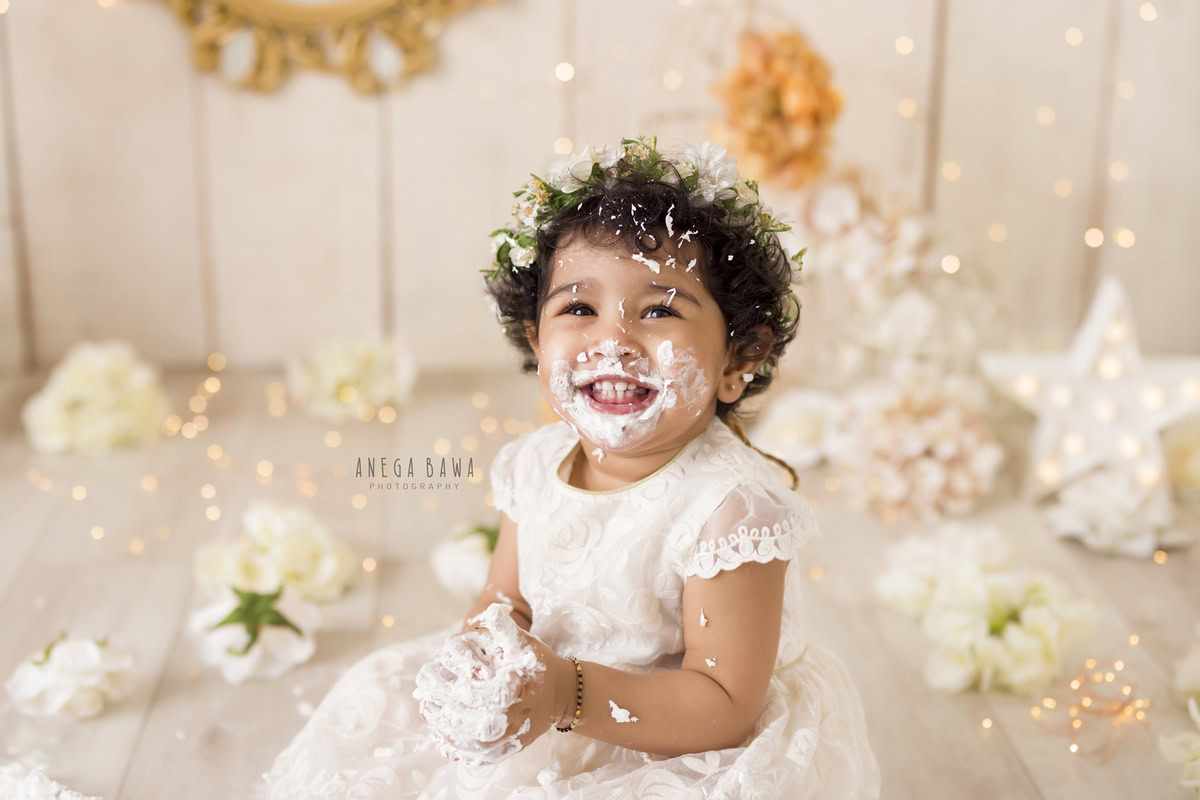 Girl with cake smash on face, white flowers, beige backdrop, fairy lights. First birthday photoshoot in Delhi, Gurgaon. Captured by Anega Bawa family photographer Gurgaon (Delhi NCR).