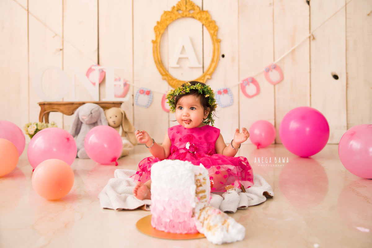 1 year old girl with cake smash surrounded by pink and peach balloons a golden frame on the wall and teddybears against a beige backdrop. Captured during a charming first birthday photoshoot in Delhi Gurgaon by the renowned family photographer Anega Bawa.