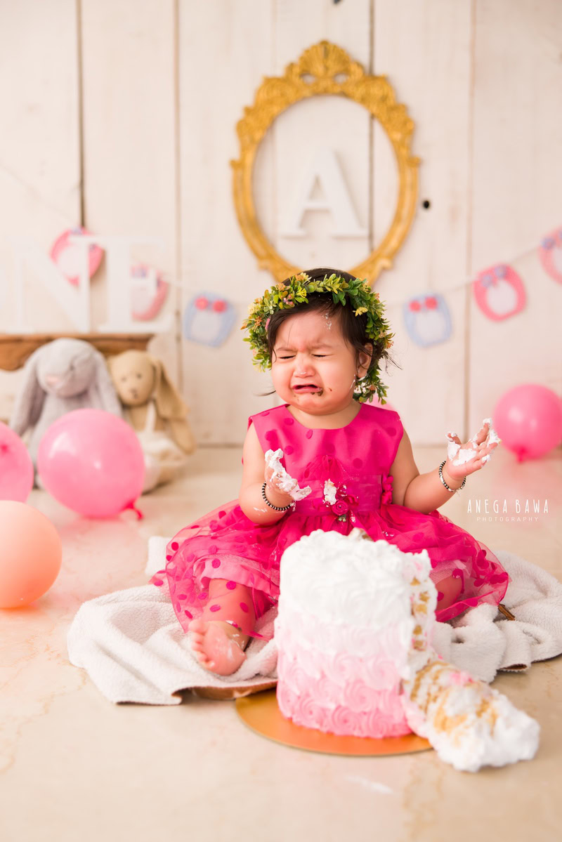 Girl with Cake Smash, wearing a tiara band, surrounded by balloons and teddy bears, against a beige backdrop, first birthday photoshoot in Delhi, Gurgaon. Captured by Anega Bawa family photographer Gurgaon (Delhi NCR)