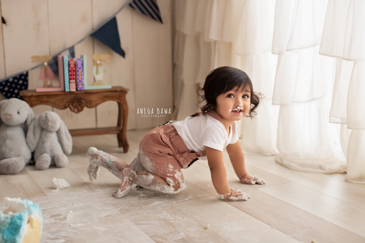 Girl crawling on the floor with Cake Smash, accompanied by a teddy bear, against a beige backdrop, first birthday photography in Delhi, Gurgaon. Captured by Anega Bawa family photographer Gurgaon (Delhi NCR)